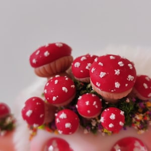 May include: A cluster of miniature red mushrooms with white spots, featuring tan stems and nestled among small patches of green moss and tiny multicolored flowers. The mushrooms are arranged in a dense group, showcasing a vibrant red and white color palette.