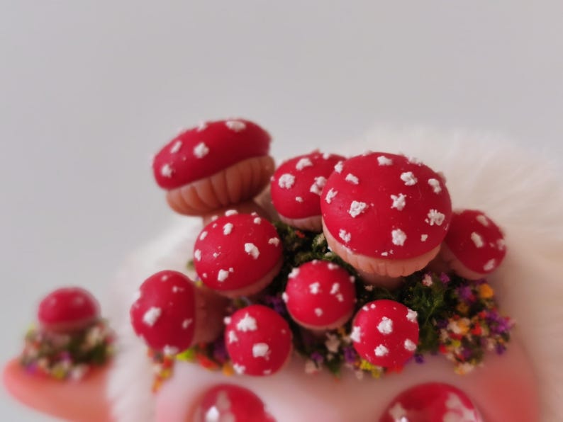 May include: A cluster of miniature red mushrooms with white spots, featuring tan stems and nestled among small patches of green moss and tiny multicolored flowers. The mushrooms are arranged in a dense group, showcasing a vibrant red and white color palette.