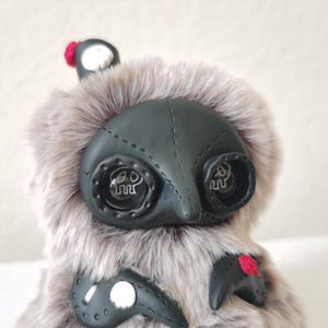 May include: A plush creature covered in long, light gray faux fur with a smooth black face mask featuring stitched details. It has two large, round, dark eyes with white skull icons inside, and small red floral accents on its body and head.