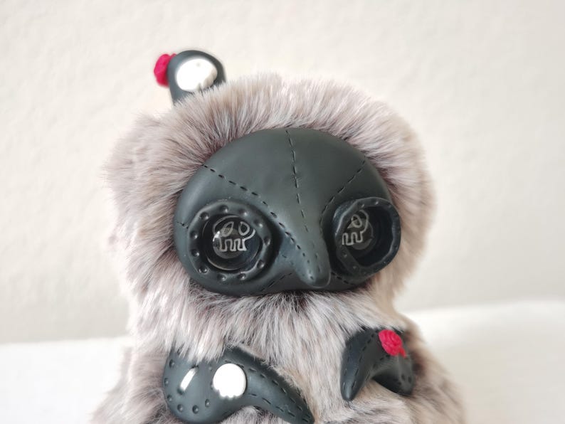 May include: A plush creature covered in long, light gray faux fur with a smooth black face mask featuring stitched details. It has two large, round, dark eyes with white skull icons inside, and small red floral accents on its body and head.