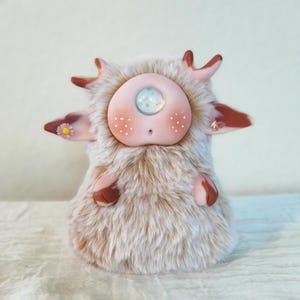 May include: A small, plush creature with beige faux fur, a single central eye, and smooth, light pink facial features. It has small antlers, two ears adorned with a tiny yellow flower and a butterfly, and two small, rounded arms with brown tips.