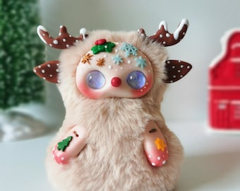 May include: A plush creature with beige faux fur, brown antlers with white spots, and a smooth face featuring large iridescent purple eyes. The face is decorated with small clay snowflakes, holly, and stars. Small green tree and yellow star accents adorn the paws.