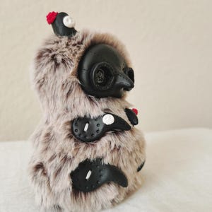 May include: A plush creature covered in long, light brown and dark brown faux fur. It features a black, mask-like face with a pointed beak, circular eye sockets, and three black, claw-like appendages on its side, each accented with a small white skull.