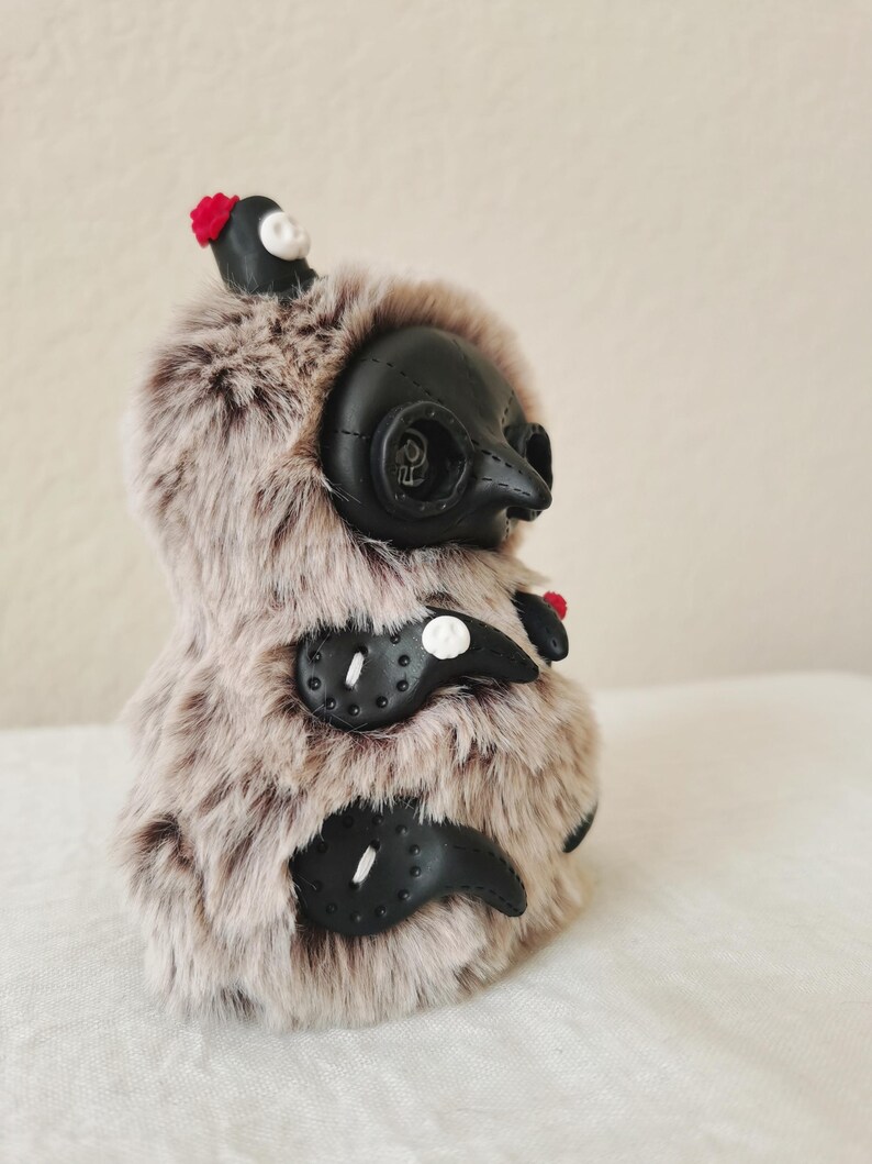 May include: A plush creature covered in long, light brown and dark brown faux fur. It features a black, mask-like face with a pointed beak, circular eye sockets, and three black, claw-like appendages on its side, each accented with a small white skull.