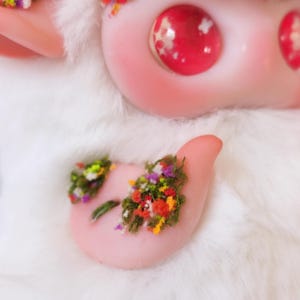 May include: A white plush creature with large, glossy, pink eyes containing white star-shaped accents. The figure features smooth, pale pink skin on the face and ears, with a small patch of green moss and tiny colorful flowers on its chest.