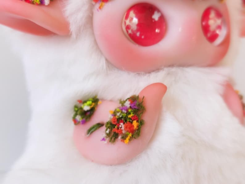 May include: A white plush creature with large, glossy, pink eyes containing white star-shaped accents. The figure features smooth, pale pink skin on the face and ears, with a small patch of green moss and tiny colorful flowers on its chest.