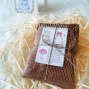 May include: A rectangular package wrapped in brown honeycomb paper, tied with thin twine. A white card featuring a red mushroom illustration and text about Gumo is attached to the front. A small matching card stands upright in the background.