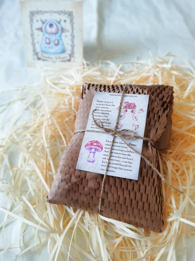 May include: A rectangular package wrapped in brown honeycomb paper, tied with thin twine. A white card featuring a red mushroom illustration and text about Gumo is attached to the front. A small matching card stands upright in the background.