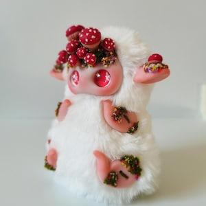 May include: A small, plush creature covered in white faux fur with large, glossy pink eyes. The figure features light pink limbs and ears, adorned with clusters of miniature red-capped mushrooms and tiny, colorful floral accents on its head and body.