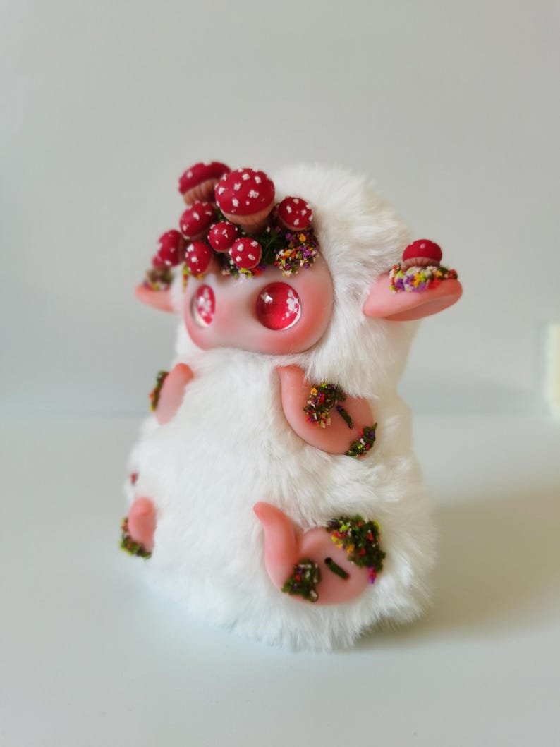 May include: A small, plush creature covered in white faux fur with large, glossy pink eyes. The figure features light pink limbs and ears, adorned with clusters of miniature red-capped mushrooms and tiny, colorful floral accents on its head and body.