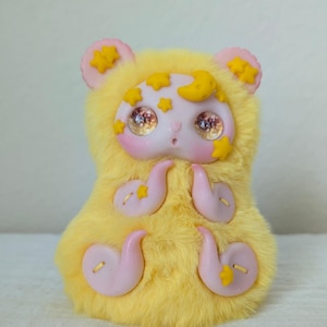 May include: A plush creature with bright yellow faux fur, featuring a smooth light pink face, large reflective brown eyes, and small pink ears. The figure is decorated with yellow stars and a crescent moon, and has four pink, curved, tentacle-like limbs.