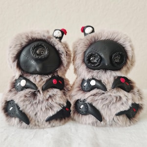 May include: Two plush creatures covered in long, light brown and grey faux fur. Each features a black face mask with button eyes, small black appendages with white or red accents, and a tiny black hat with a red or white skull detail on top.
