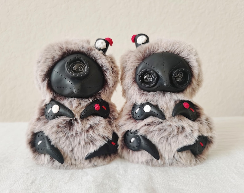 May include: Two plush creatures covered in long, light brown and grey faux fur. Each features a black face mask with button eyes, small black appendages with white or red accents, and a tiny black hat with a red or white skull detail on top.