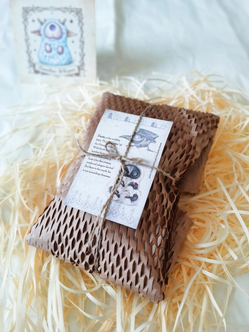 May include: A rectangular package wrapped in brown honeycomb paper, secured with thin twine. A white rectangular card is tied to the front, featuring a black and white illustration of a plague doctor and text describing Dusty the wobbly little doctor.