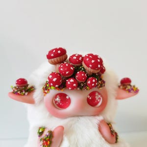 May include: A white plush creature with large, glossy pink eyes and a cluster of red mushrooms with white spots on its head. The creature features small pink ears and arms decorated with tiny, colorful floral accents and green foliage details.