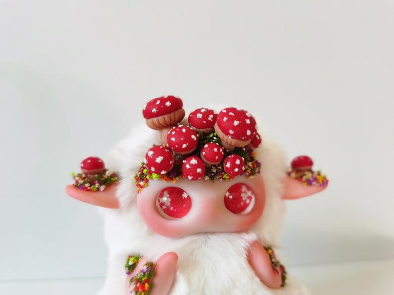 May include: A white plush creature with large, glossy pink eyes and a cluster of red mushrooms with white spots on its head. The creature features small pink ears and arms decorated with tiny, colorful floral accents and green foliage details.