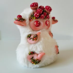 May include: A plush creature covered in white faux fur with pink skin accents on the ears, hands, and feet. The figure features red button eyes and is adorned with clusters of small red mushrooms with white spots and patches of colorful floral moss.