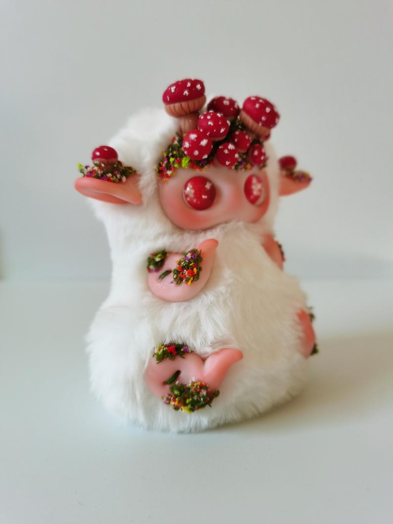 May include: A plush creature covered in white faux fur with pink skin accents on the ears, hands, and feet. The figure features red button eyes and is adorned with clusters of small red mushrooms with white spots and patches of colorful floral moss.
