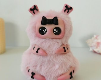 May include: A small, plush, light pink creature figurine with a smooth face, large black eyes, and a black bow on its head. It features fuzzy pink fur, small pink limbs with black bow accents, and two small ears, each decorated with a black bow.