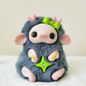 May include: A plush creature with dark blue faux fur, light pink felt ears, antlers, and paws. It features large black eyes, a light pink face, a bright green bow on its head, and a four-pointed star-shaped green button on its chest.