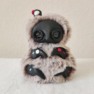 May include: A plush creature covered in long, light brown faux fur with a black, mask-like face featuring two large, dark, circular eyes with skull-shaped pupils. It has four black, claw-like appendages adorned with small white and red accents.