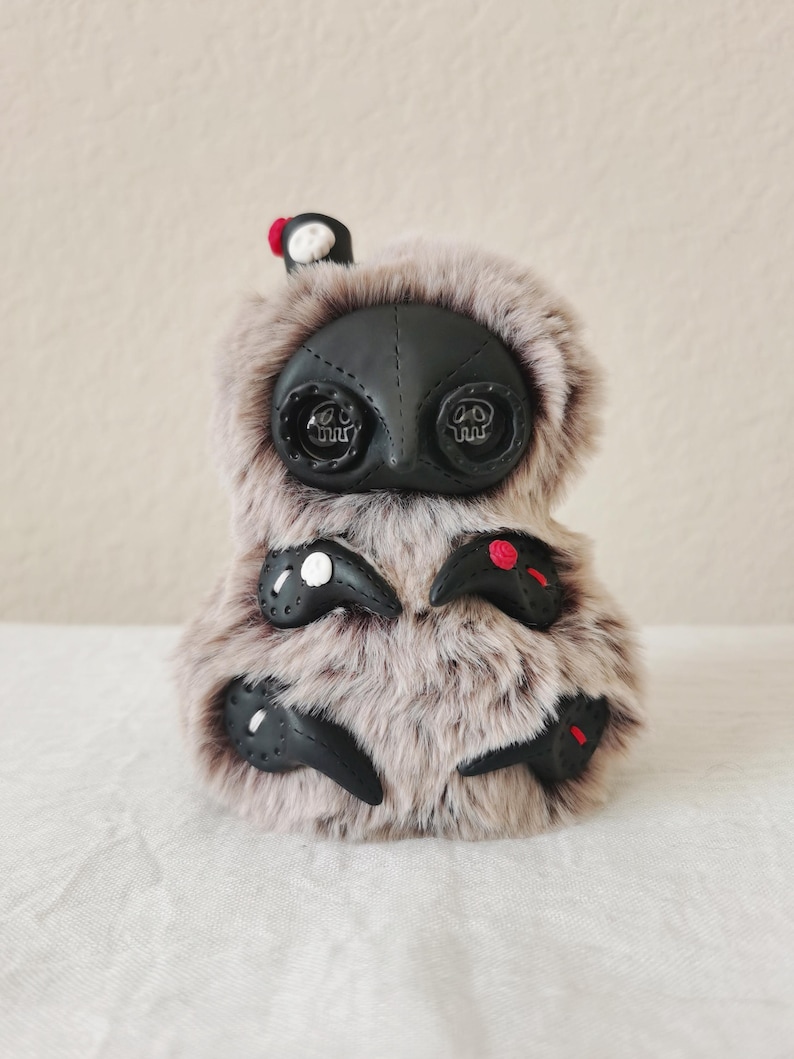May include: A plush creature covered in long, light brown faux fur with a black, mask-like face featuring two large, dark, circular eyes with skull-shaped pupils. It has four black, claw-like appendages adorned with small white and red accents.