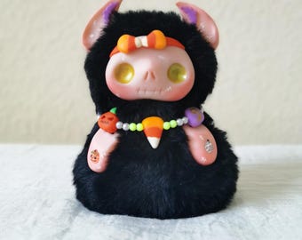 May include: A small, plush creature with black fur, a pale pink face, and yellow eyes. It wears a candy corn-themed headband and a beaded necklace featuring a candy corn charm, a small orange pumpkin, and a purple accent piece on its neck.