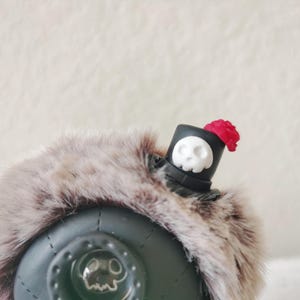 May include: A dark green spherical object adorned with a thick, brown and gray faux fur trim. A small black top hat sits on the fur, featuring a white skull emblem on the front and a small red rose accent on the side. A skull icon is visible.