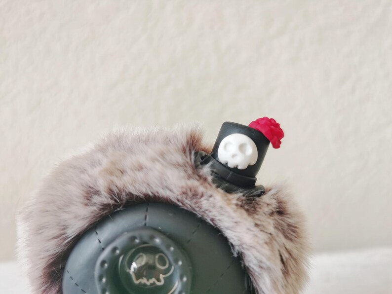 May include: A dark green spherical object adorned with a thick, brown and gray faux fur trim. A small black top hat sits on the fur, featuring a white skull emblem on the front and a small red rose accent on the side. A skull icon is visible.
