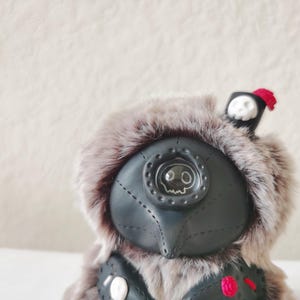 May include: A plush creature with grey faux fur and a black leather-like mask featuring a single central eye with a skull graphic. The figure wears a small black hat with a white skull emblem and a red tassel, plus a black vest with red accents.