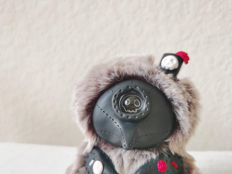 May include: A plush creature with grey faux fur and a black leather-like mask featuring a single central eye with a skull graphic. The figure wears a small black hat with a white skull emblem and a red tassel, plus a black vest with red accents.