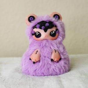 May include: A small creature figurine covered in soft lavender faux fur. It features a smooth, pale peach-colored face with large, dark, glossy eyes, black clawed hands, and a headpiece adorned with black and purple petal-like accents.