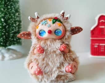 May include: A small, fuzzy, tan-colored creature figurine with large, round, blue eyes and a red nose. It features antlers, small ears with white dots, and decorative accents on its forehead including snowflakes, stars, and holly leaves.