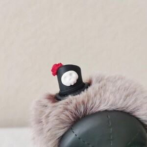 May include: A dark green beret with a thick, light brown faux fur trim. Attached to the top is a small black top hat ornament featuring a white skull emblem on the front and a tiny red flower accent on the side of the hat brim.