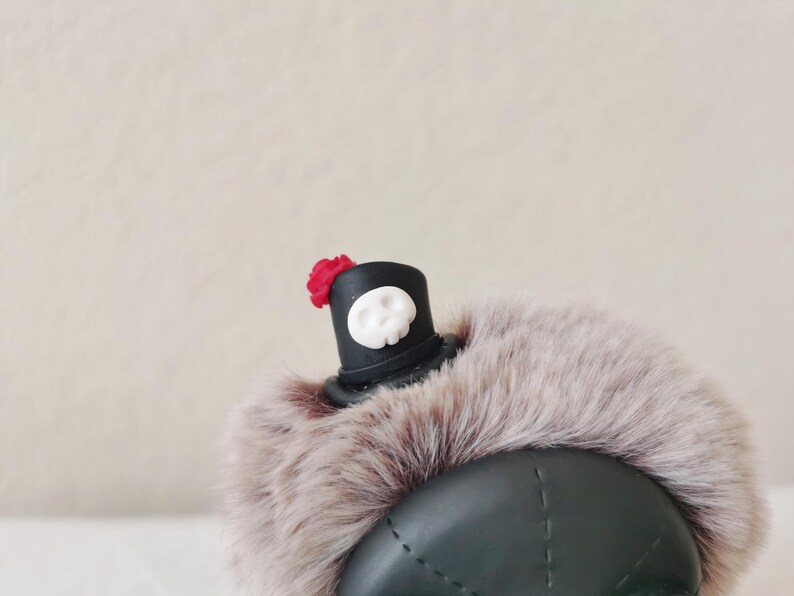 May include: A dark green beret with a thick, light brown faux fur trim. Attached to the top is a small black top hat ornament featuring a white skull emblem on the front and a tiny red flower accent on the side of the hat brim.