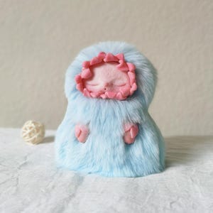 May include: A small figurine featuring a pale pink face with closed eyes, framed by a ruffled pink border, all encased in a soft, light blue faux fur body. Two small pink hands protrude from the fur, and a small white woven sphere sits nearby.