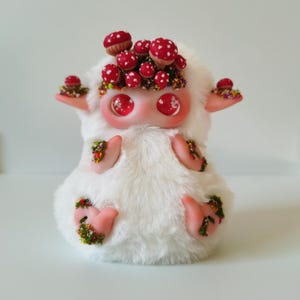 May include: A white plush creature with large glossy red eyes and pointed ears. The figure is adorned with numerous small red mushrooms with white spots and patches of colorful moss on its head, ears, hands, and feet, creating a textured appearance.