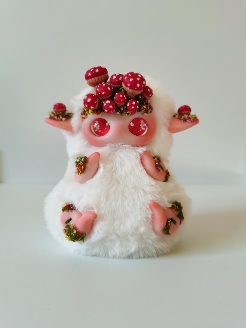 May include: A white plush creature with large glossy red eyes and pointed ears. The figure is adorned with numerous small red mushrooms with white spots and patches of colorful moss on its head, ears, hands, and feet, creating a textured appearance.