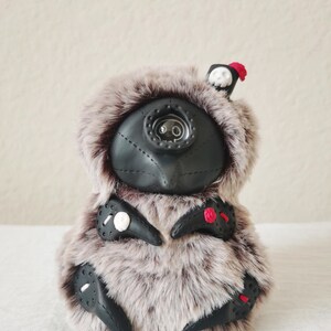 May include: A plush creature with long, light brown and grey faux fur, featuring a black, mask-like face with a central eye-like detail. It has black, claw-like appendages adorned with small white skulls, a red rose, and red accents.