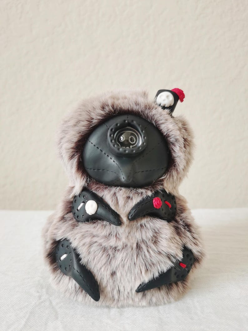 May include: A plush creature with long, light brown and grey faux fur, featuring a black, mask-like face with a central eye-like detail. It has black, claw-like appendages adorned with small white skulls, a red rose, and red accents.