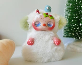 May include: A small, plush creature with white faux fur, light green ear-like tufts, and pink polka-dot hands. The face features large, iridescent blue eyes, a small orange nose, and a festive headpiece with a tiny snowman and holly.