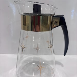 Puede incluir: Una jarra de cristal transparente con una banda de color dorado alrededor de la parte superior y un asa negra. El cuerpo de la jarra está decorado con un patrón de estrellas doradas. Incluye una tapa negra con un pomo central.