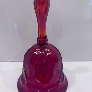 May include: A deep ruby red glass bell featuring a faceted geometric pattern on the main body and a smooth, elongated handle. The translucent material catches the light, highlighting the sharp edges of the cut glass design and the flared base.