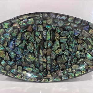 May include: An oval-shaped decorative tray featuring a mosaic surface composed of numerous small, iridescent abalone shell fragments in shades of green, blue, and purple, set against a dark background with a smooth, glossy finish.