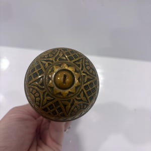 May include: A round brass door knob featuring an intricate embossed geometric and floral pattern across its surface. The centre of the knob displays a circular plate with a sunburst edge surrounding a vertical keyhole slot for a locking mechanism.