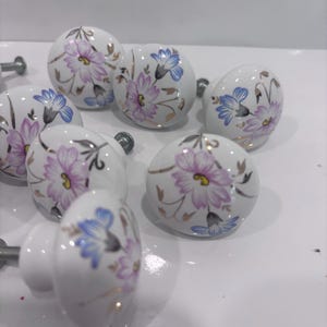 May include: A collection of seven round white ceramic cabinet knobs, each featuring a hand-painted floral design with light purple and blue petals, yellow centers, and delicate gold-toned stems and leaves, complete with metal mounting screws.