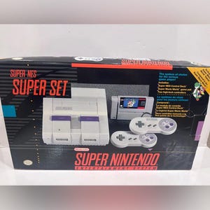 May include: A Super Nintendo Entertainment System Super Set box featuring a grey game console, two grey controllers, and a Super Mario World game cartridge. The black packaging displays the Super NES logo, product text, and a graphic of the console.