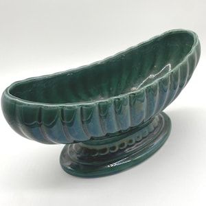 May include: A vintage, oval-shaped, green ceramic planter with a pedestal base. The planter has a ribbed exterior and a glossy finish. The base is oval with a decorative beaded design. The planter is suitable for flowers or other decorative plants.