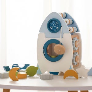 May include: A wooden toy rocket ship in white and light blue with a porthole and a small clock face. The rocket has wooden gears and beads, and is surrounded by colorful wooden shapes, including a star, moon, and cloud.