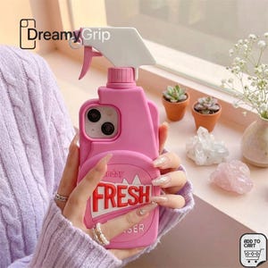 May include: A pink silicone phone case shaped like a spray bottle with a white trigger nozzle at the top. The case features the word FRESH in bold red letters on the front, with a cutout for the camera lens and a pink cap detail.